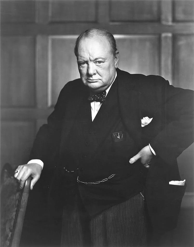 Das Bild zeigt Winston Churchill im Anzug mit einer Fliege und Taschenuhrkette, der sich an einen Stuhl lehnt. Er blickt ernst in die Kamera und trägt eine gepunktete Fliege sowie ein Einstecktuch.