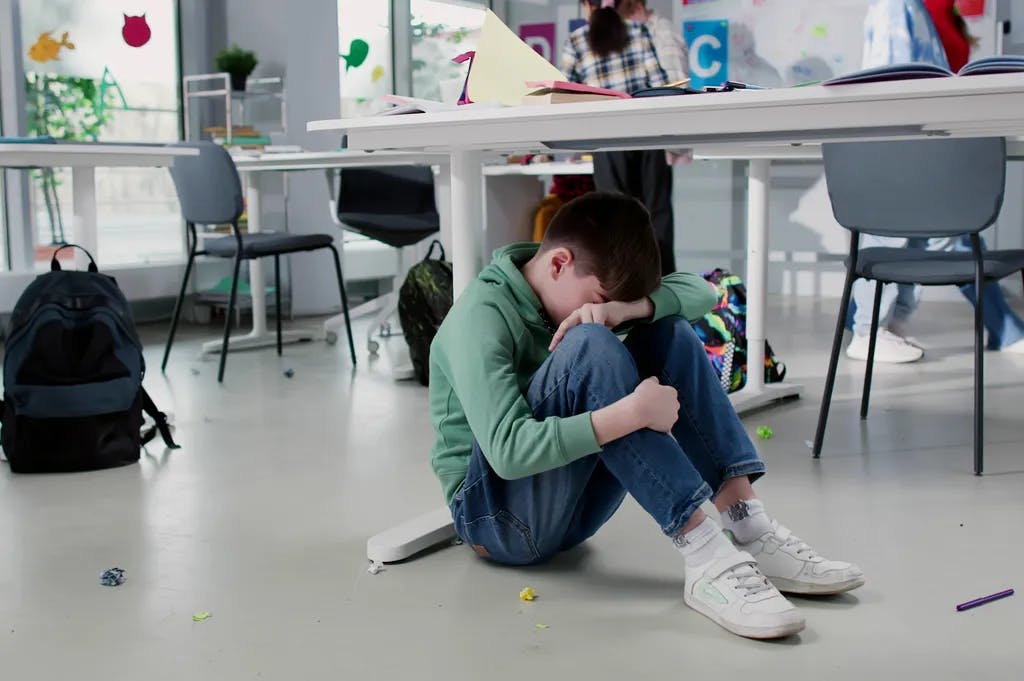 Depressiver einsamer Junge, der unter einem Schreibtisch im Klassenzimmer sitzt und auf dem Hintergrund spielt
