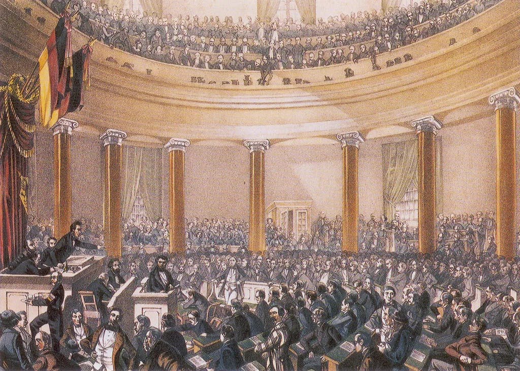 Im Rahmen der Deutschen Revolution 1848/49 zeigt dieses Bild die Nationalversammlung in der Paulskirche in Frankfurt. Eine große Menge von Delegierten und Zuschauern ist in einem halbrunden Saal versammelt, einige stehen und es wird eine Rede gehalten.
