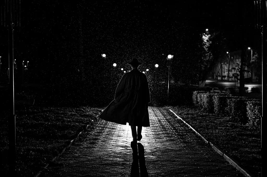 dunkle Silhouette eines männlichen Detektivs in einem Mantel und Hut im Regen in einer Nachtstraße im Stil Noir